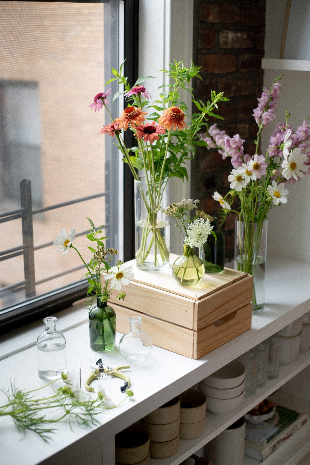 A styled arrangement of fresh cut flowers in clear glass vases and a small green bottle, displayed on a white shelf next to a window with a brick wall in the background. A light wood box sits beneath the vases.